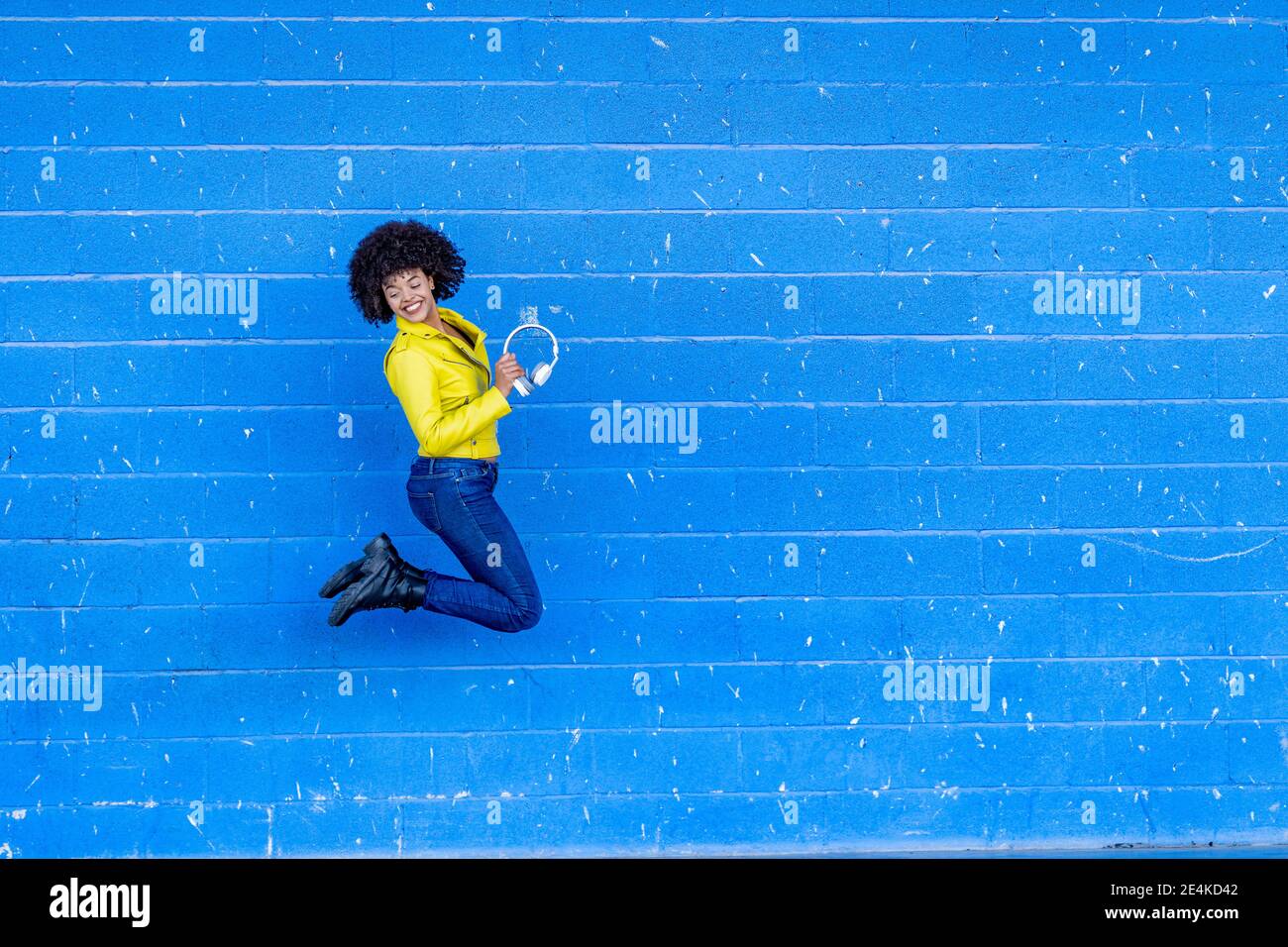 Fröhliche junge Frau hält Kopfhörer, während sie gegen die blaue Wand springt Stockfoto