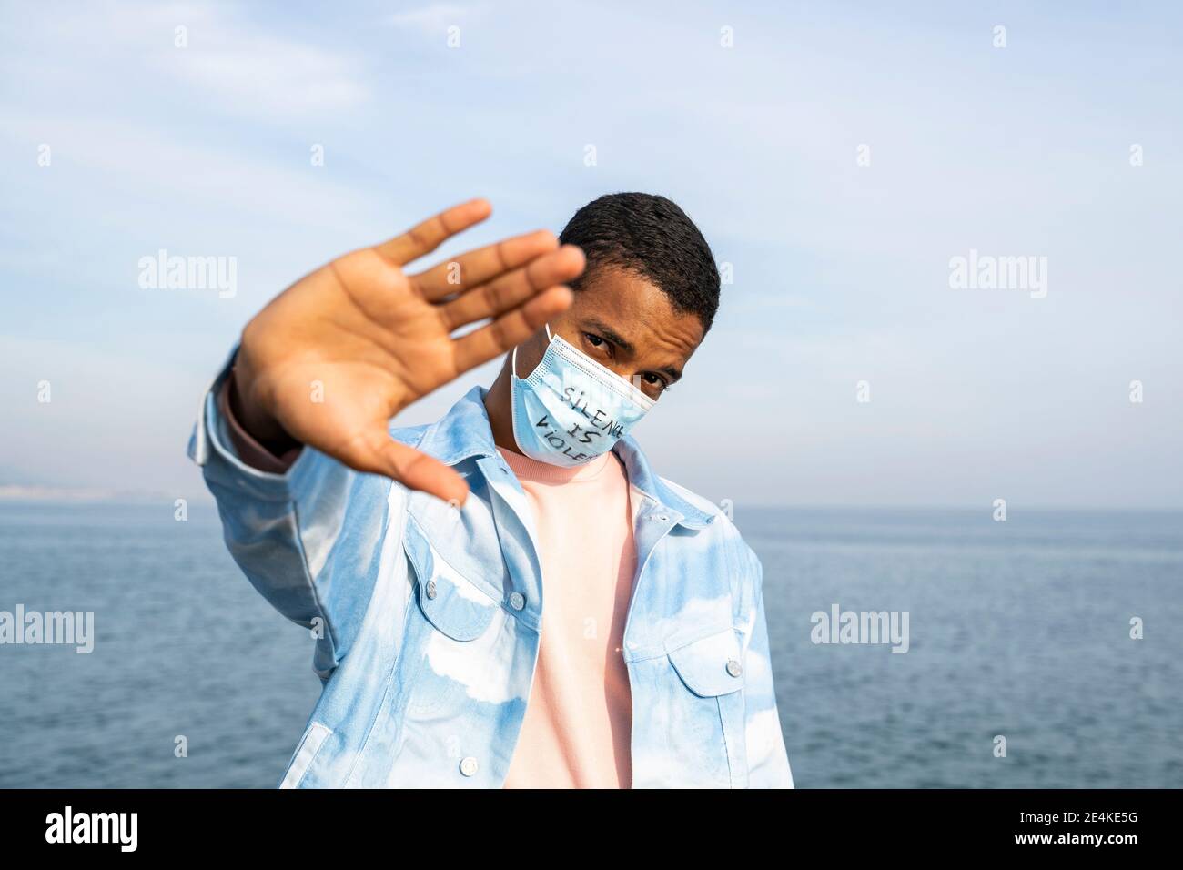 Junger Mann mit der Gesichtsmaske „Silence is Violence“, die zeigt Stop-Geste, während Sie gegen das Meer stehen Stockfoto