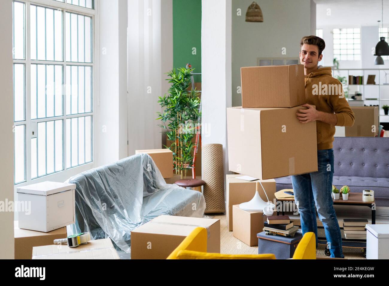 Lächelnder junger Mann, der Pappkartons im Wohnzimmer trägt Neue Wohnung Stockfoto