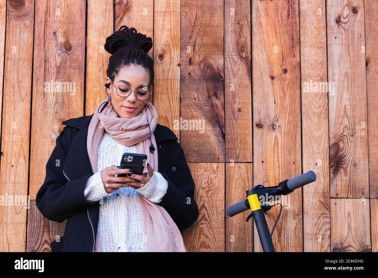 Junge Frau Millennial mit Smartphone, während lehnte sich gegen Holz Wand durch elektrische Push-Roller Stockfoto