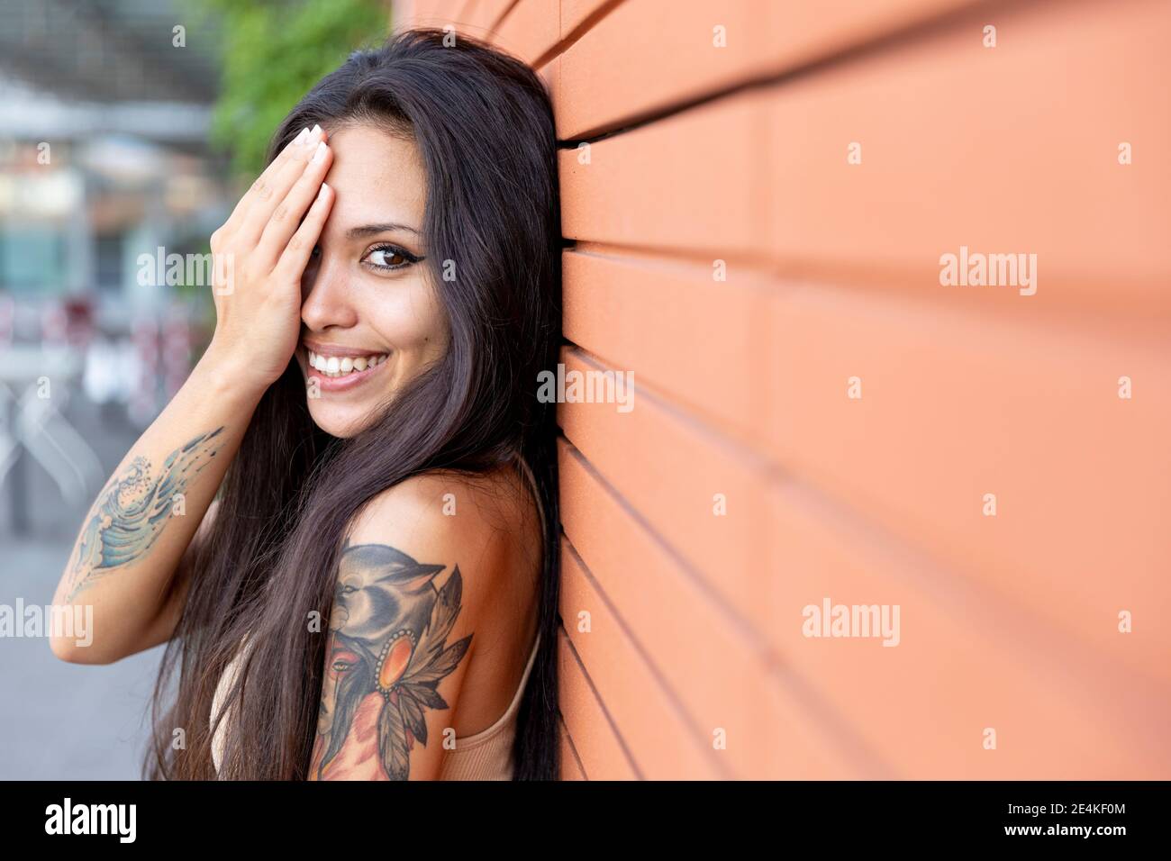 Junge Frau lächelt, während sie das Gesicht mit der Hand bedeckt, die sich darauf stützt Wand Stockfoto