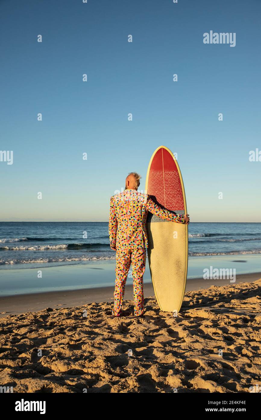 Mann im Anzug hält Surfbrett, während er am Strand steht Stockfoto