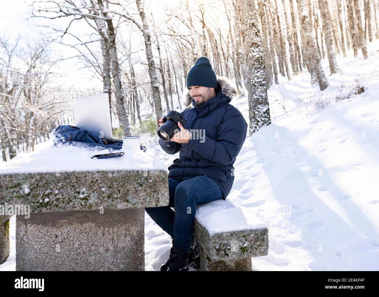 Lächelnder Fotograf mit Kamera, während er auf einer Felsbank sitzt Wald bei schneebedecktem Wetter Stockfoto