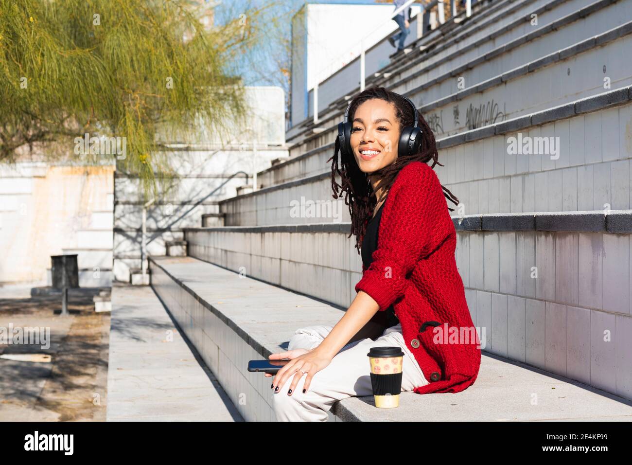 Lächelnd junge weibliche Millennial sitzen mit Smartphone und wiederverwendbar pokal auf der Treppe an sonnigen Tag Stockfoto