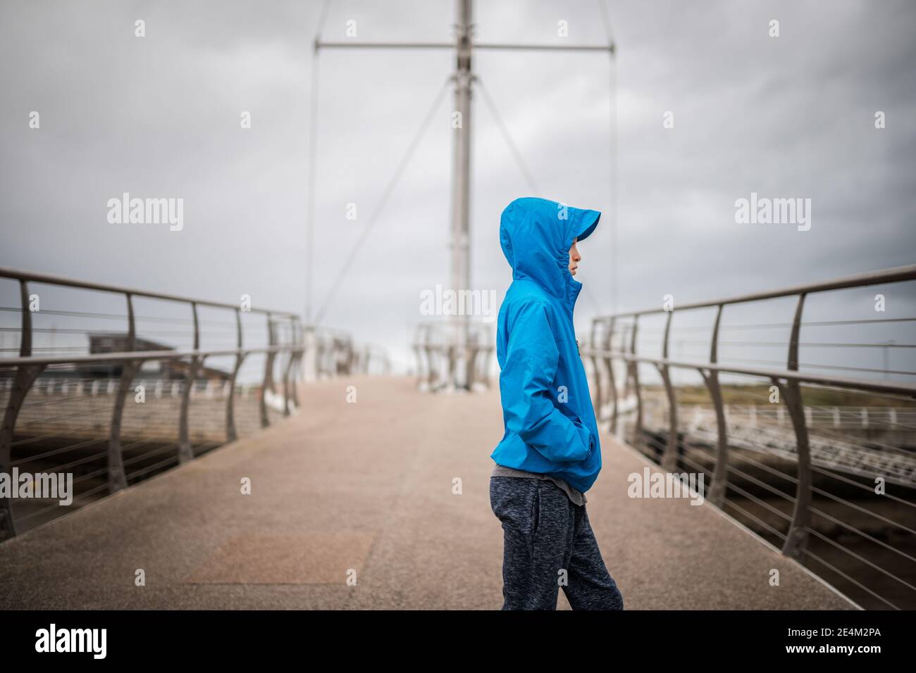 Junge in blauem Mantel mit Kapuze auf Brücke suchen Weg von der Kamera Seite Regenmantel Metall Geländer Boot Segel Hintergrund Hafen Ozean Hände in Taschen kalt Stockfoto