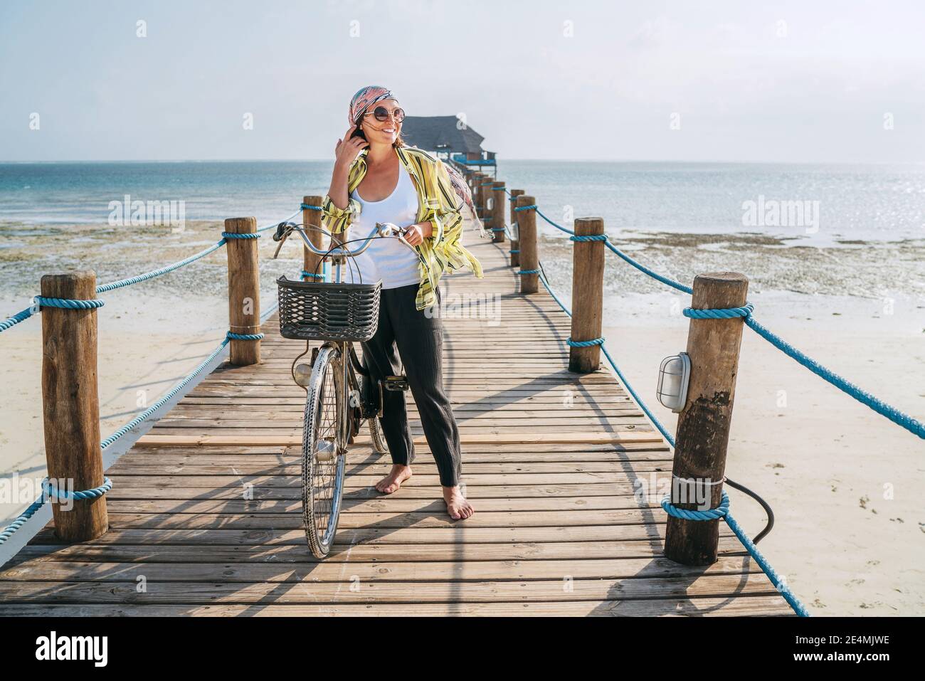 Porträt einer lächelnden Frau in leichte Sommerkleidung gekleidet, Sonnenbrille mit Fahrrad auf dem hölzernen Seebrücke am Sandstrand von Sansibar.sorgloser Urlaub Stockfoto