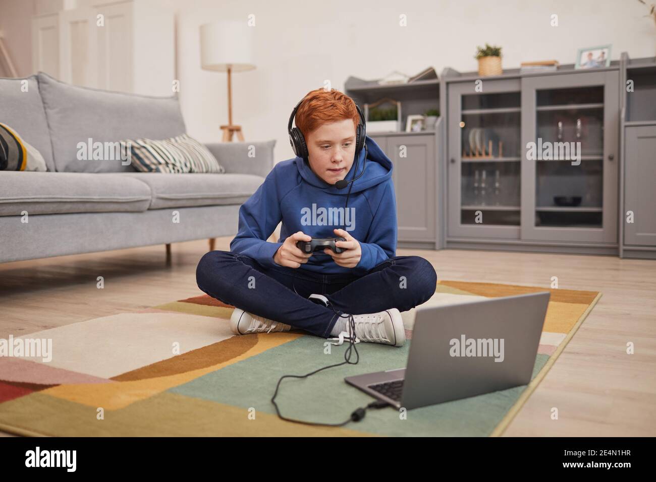 In voller Länge Porträt von rothaarigen Teenager spielen Videospiele, während auf dem Boden sitzen und halten Gamepad, kopieren Raum Stockfoto
