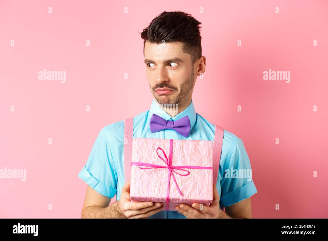 Feiertags- und Feiertagskonzept. Schockiert Kerl neben starrte mit Cringe Gesicht, hält kleine Geschenkbox, stand über rosa Hintergrund Stockfoto