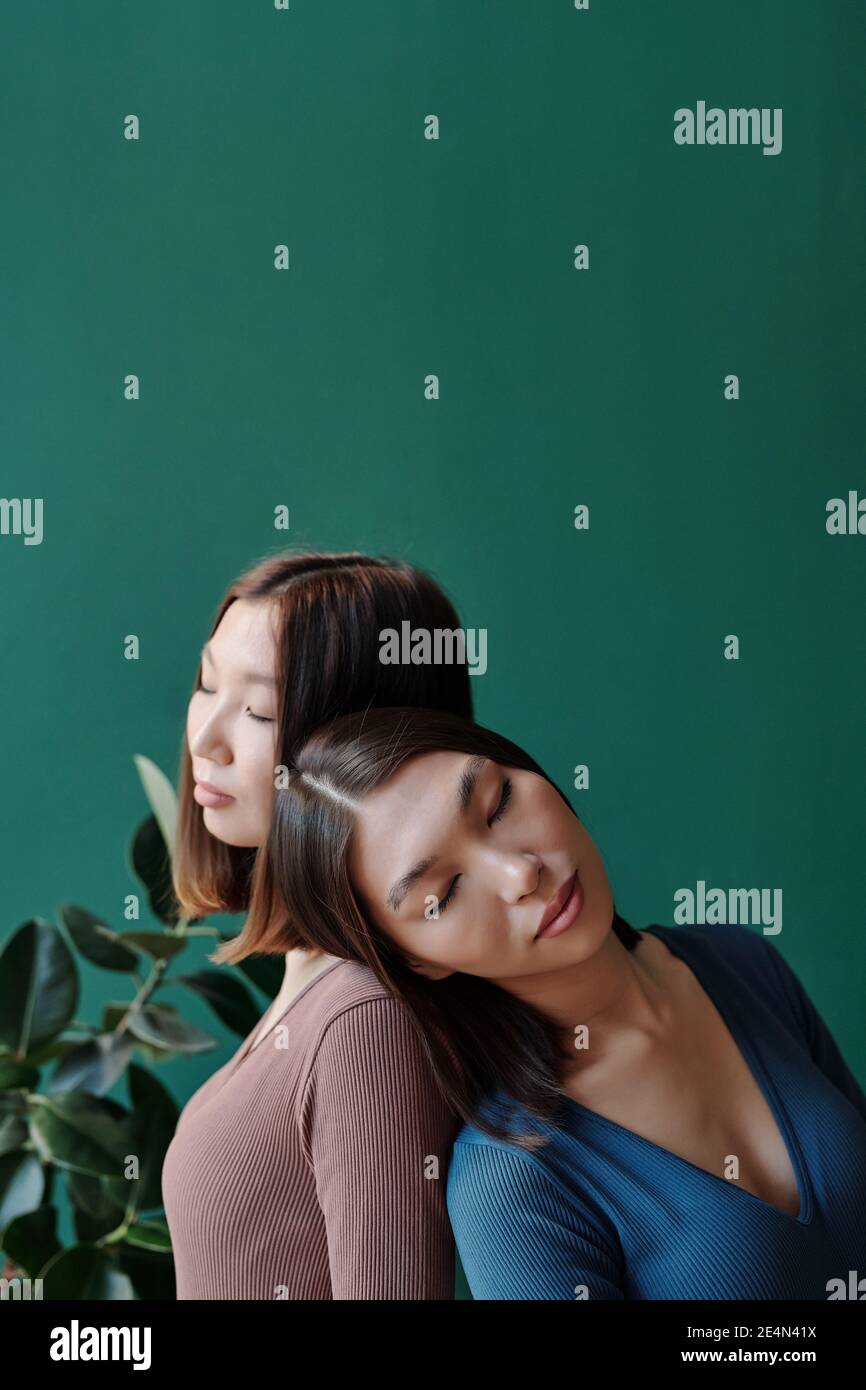 Junge Zwillingsschwestern asiatischer Ethnie mit dunklem, glattem Haar Entspannen vor der Kamera gegen Hausanlage mit Grün Blätter an Jadewand Stockfoto