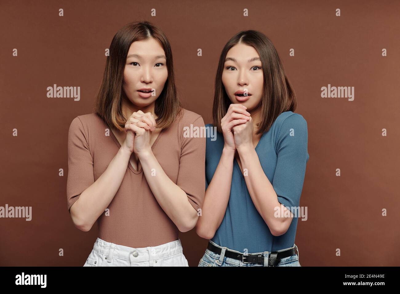 Zwei junge erstaunt Brünette Frauen der asiatischen Ethnie halten Hände Zusammen und gekreuzt von Chins, während Ausdruck großer Erstaunen Stockfoto