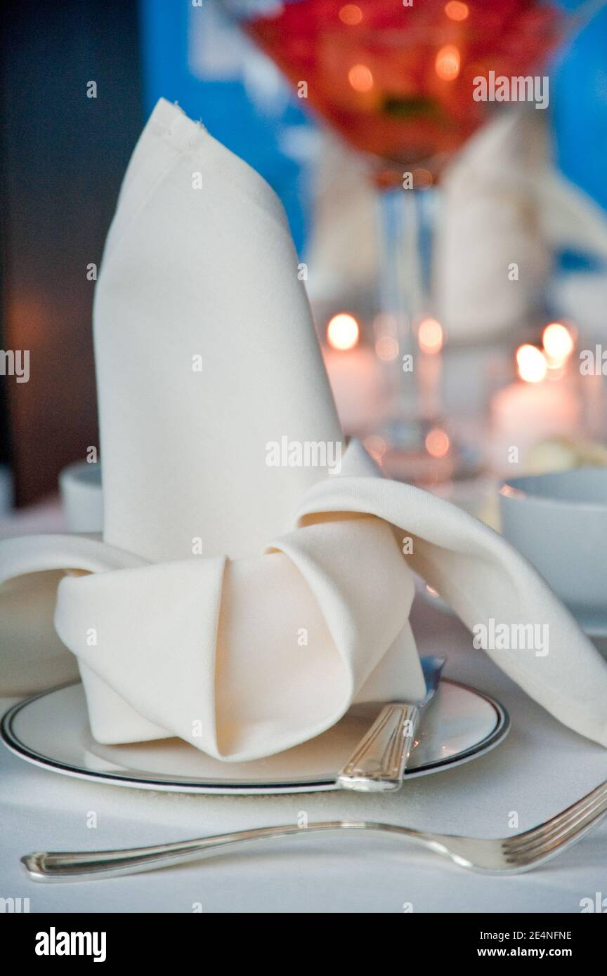 Weiße gefaltete Serviette auf feinen Dining-Tellern mit Cocktailglas Und Kerzen im Hintergrund Stockfoto