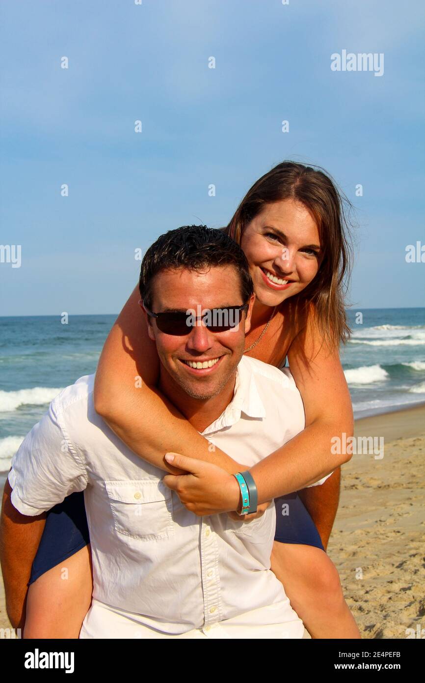 Ein brünette kaukasisches Paar genießt eine Huckepack Fahrt mit Meer Im Hintergrund Stockfoto