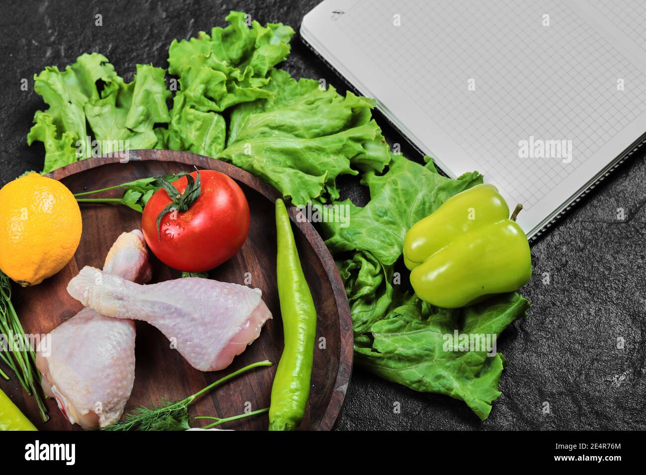 Rohe Hähnchenbeine auf Holzplatte mit frischem Gemüse und Öffnen Sie das Notizbuch Stockfoto