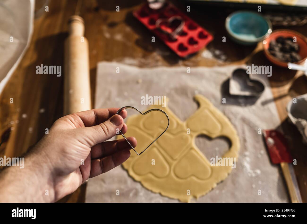 Mann hält Eisenkuchen-Kuchenform mit Herz in der Hand. Rohe Teigkekse auf dem Tisch machen. Stockfoto
