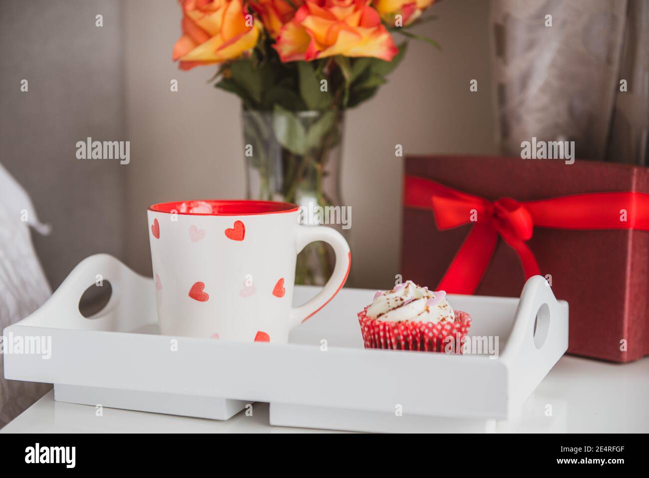 Tablett mit frischem Kaffee und Cupcake, Geschenkbox und einem Strauß Rosenblumen in Vase auf dem Nachttisch gegen eine graue Wand. Valentinstag Stockfoto