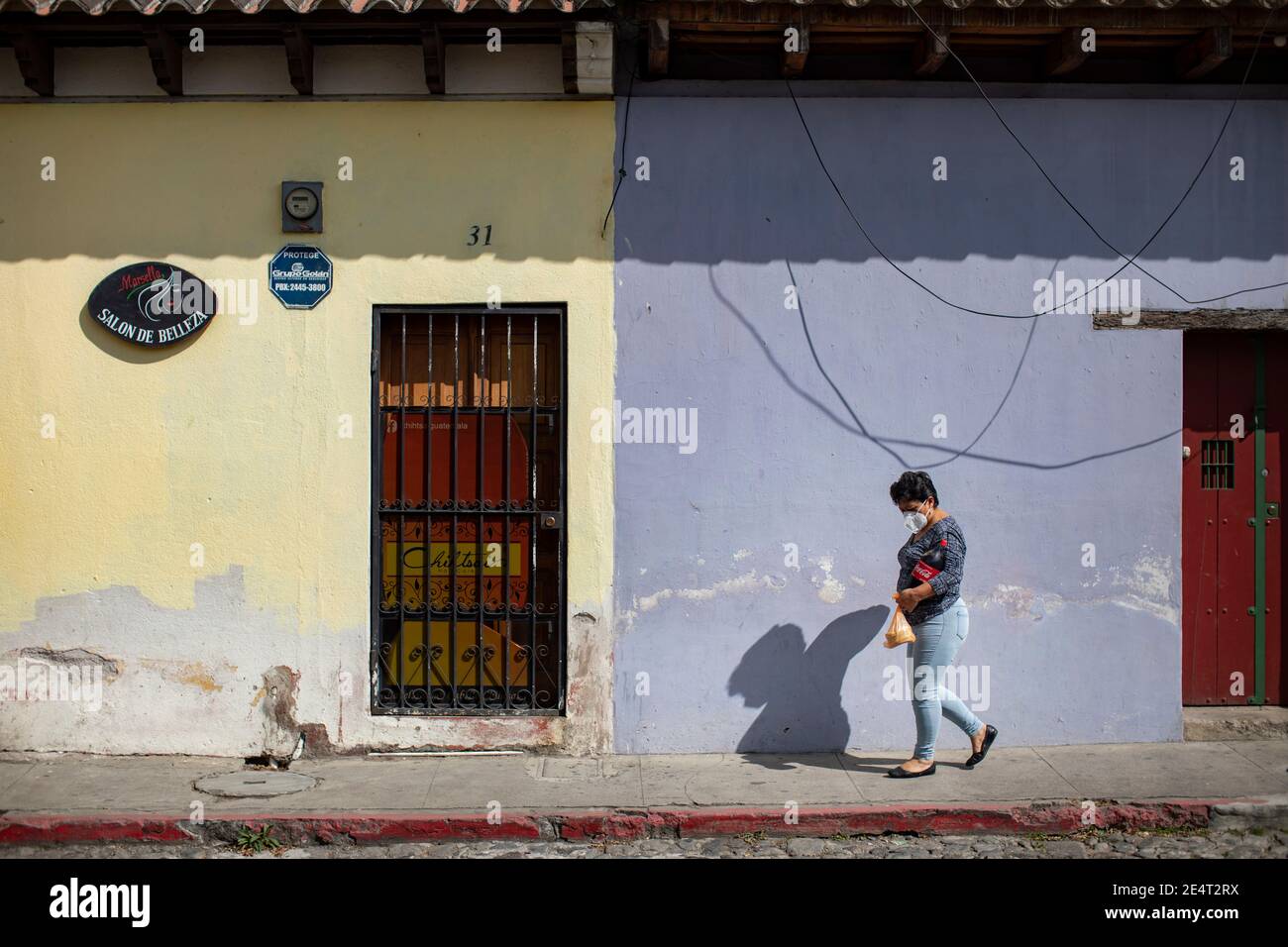 Frau mit Maske auf dem Bürgersteig vor bunten Häusern und Geschäften in Antigua, Guatemala, Mittelamerika. Stockfoto