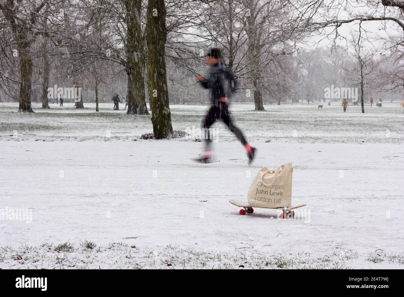 Mann, der an einem Skateboard mit John Lewis entlang läuft Einkaufstasche bei schwerem schneebedeckten Winterwetter in London Greenwich parken Stockfoto