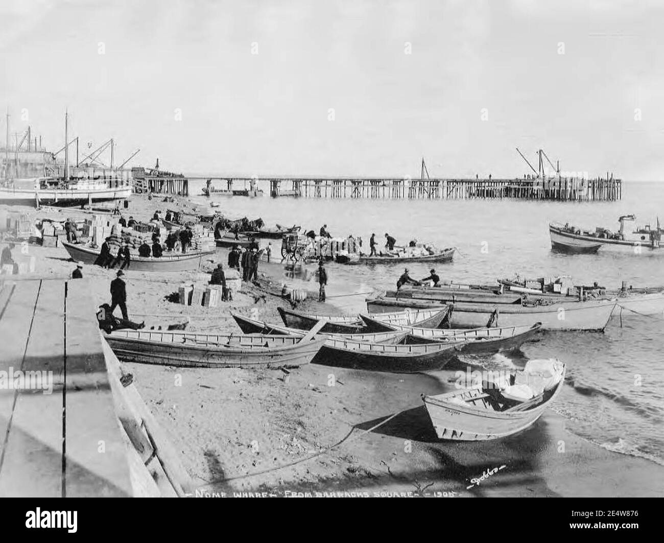 Männer, die Ladung von kleinen Booten am Strand entladen, mit Anlegestelle (wahrscheinlich Sesnon Wharf) im Hintergrund, Nome, 1905 (AL+CA 6518). Stockfoto