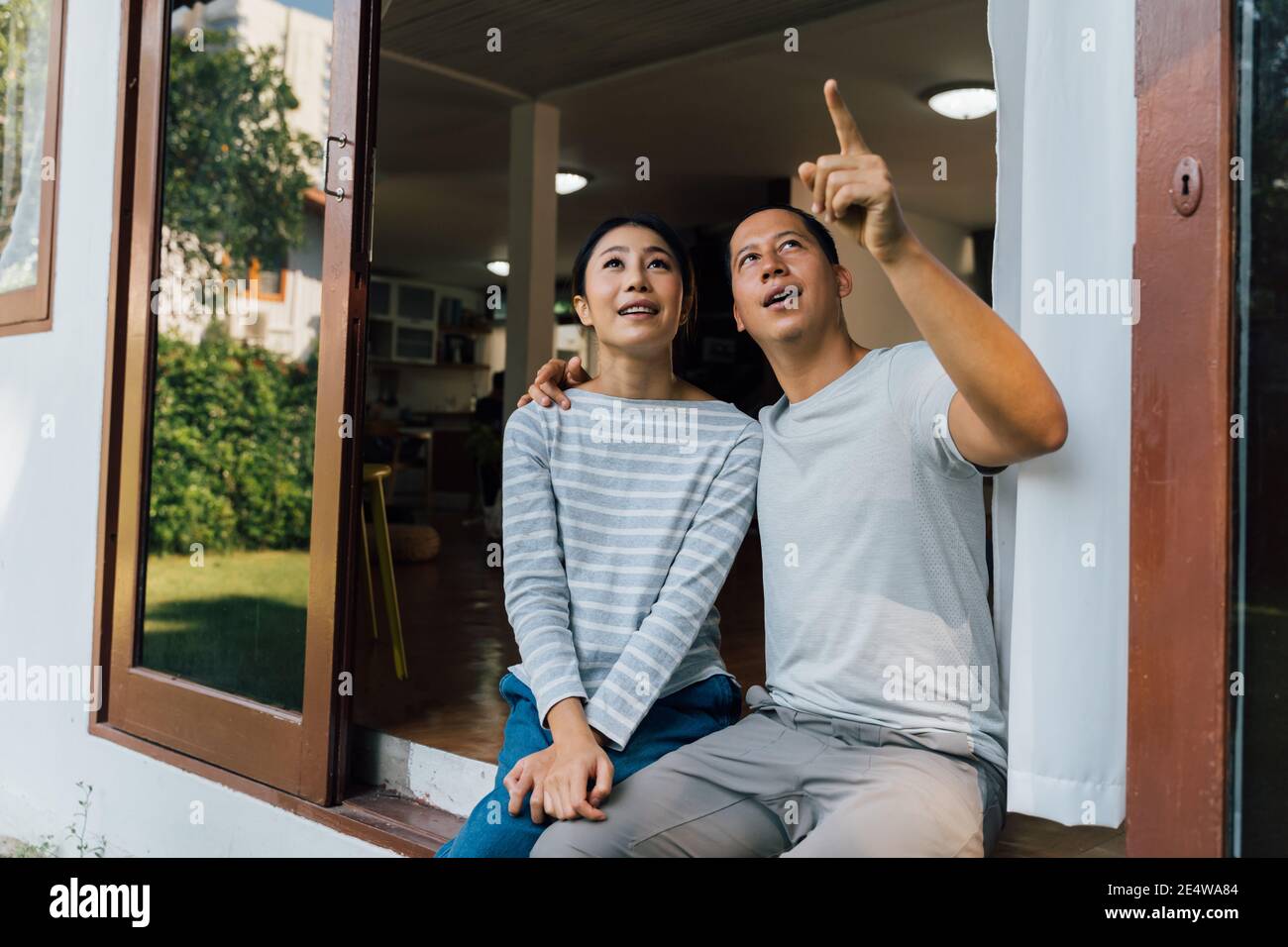Portrait von jungen erwachsenen asiatischen Paar suchen nach oben, während ein Mann zeigt mit Finger in Hause inneren Hintergrund. 30s reifen erfolgreichen Ehemann und Ehefrau. Immobilien- und Immobilienleistungskonzept Stockfoto