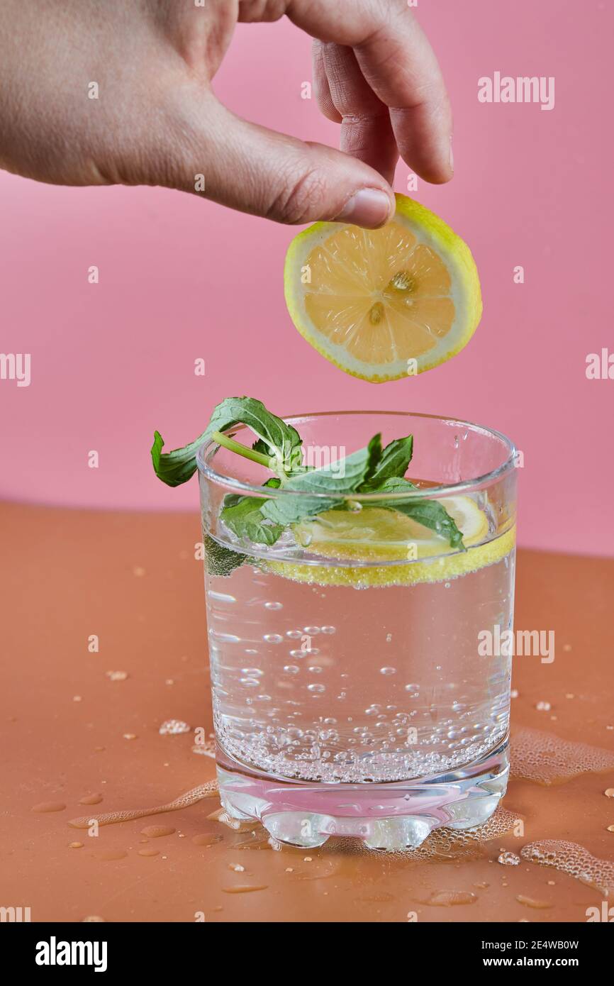 Ein Glas kalte erfrischende Limonade auf rosa Hintergrund und Frau hält eine Zitronenscheibe Stockfoto