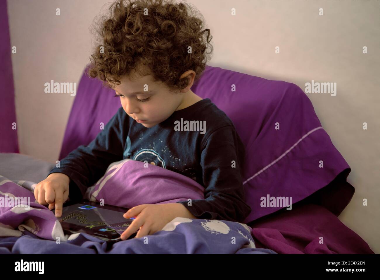 Sweet 4-5 Jahre alten Jungen mit lockigen Haaren im Bett wach in den Morgen, beobachten Cartoons auf Tablet, Unterhaltung sich drinnen. Neues normales Konzept Stockfoto