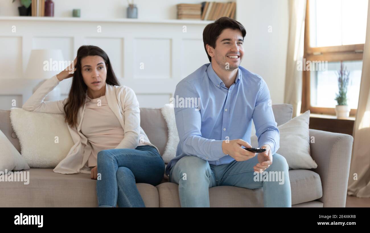 Lächelnder Mann beim Fernsehen, gelangweilte wütende Frau, die getrennt sitzt Stockfoto