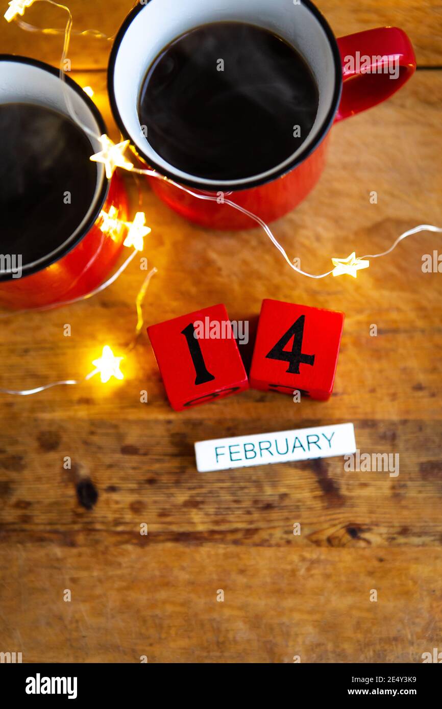 Zwei Tassen Kaffee in roten Tassen auf einem Hintergrund von Girlanden. Postkarte mit Datum und Monat. Frühstück am Valentinstag. Blick von oben Stockfoto