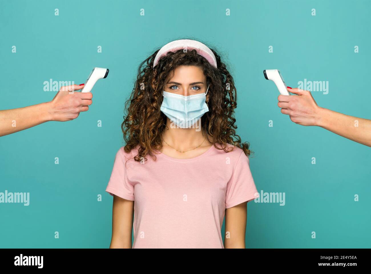 Junge Frau in schützende Gesichtsmaske während der Covid-19 oder Coronavirus-Pandemie, deren Temperatur auf einem digitalen Thermometer überwacht wird Durch zwei Arme Stockfoto