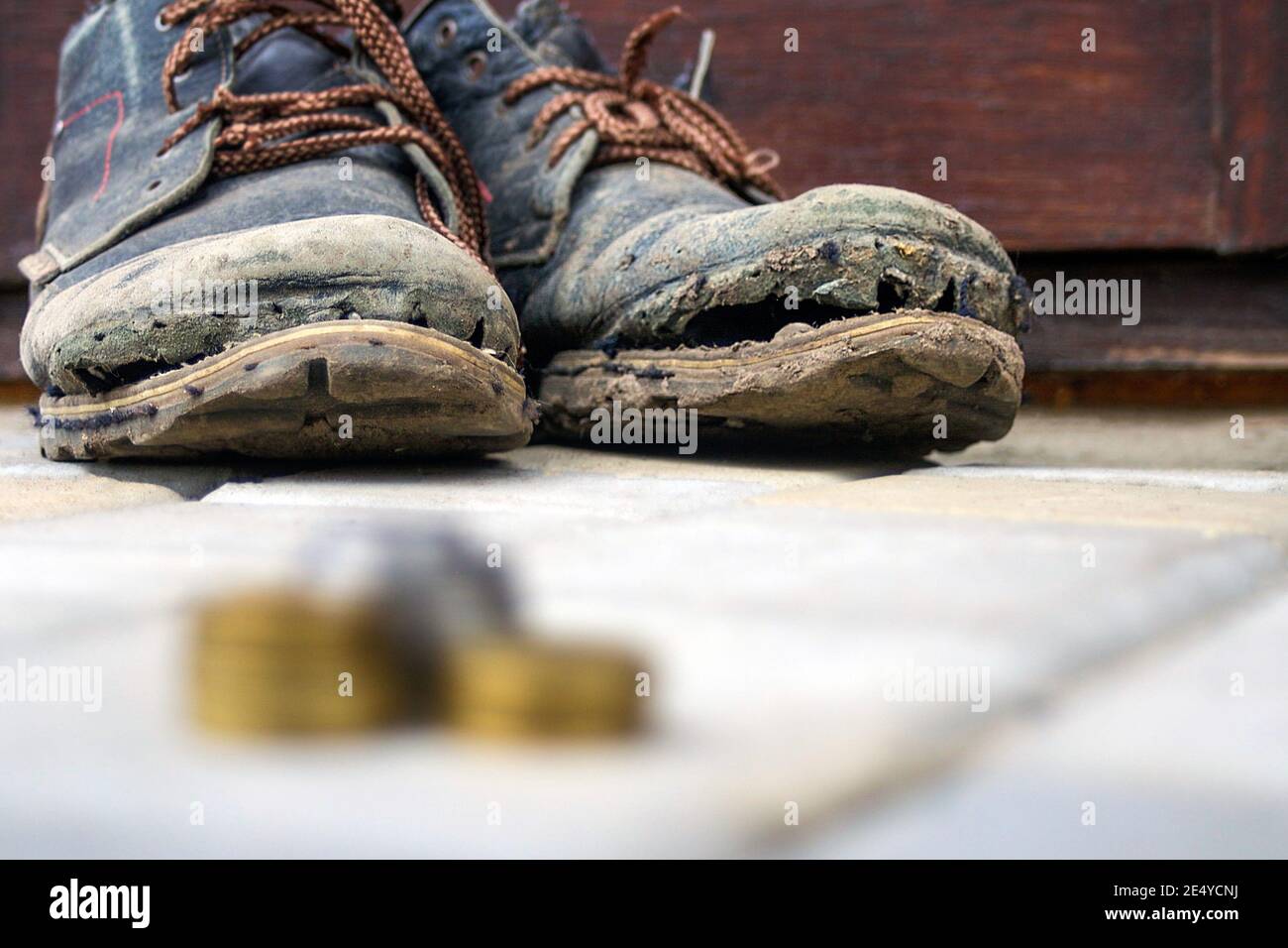 Abgenutzte Stiefel mit einer nachlagenden Sohle auf einem Hintergrund aus grauen Fertigern und dunkelbraunem Holz. Im Vordergrund ist ein verschwommener Stapel von Münzen. Das Konzept des Povers Stockfoto