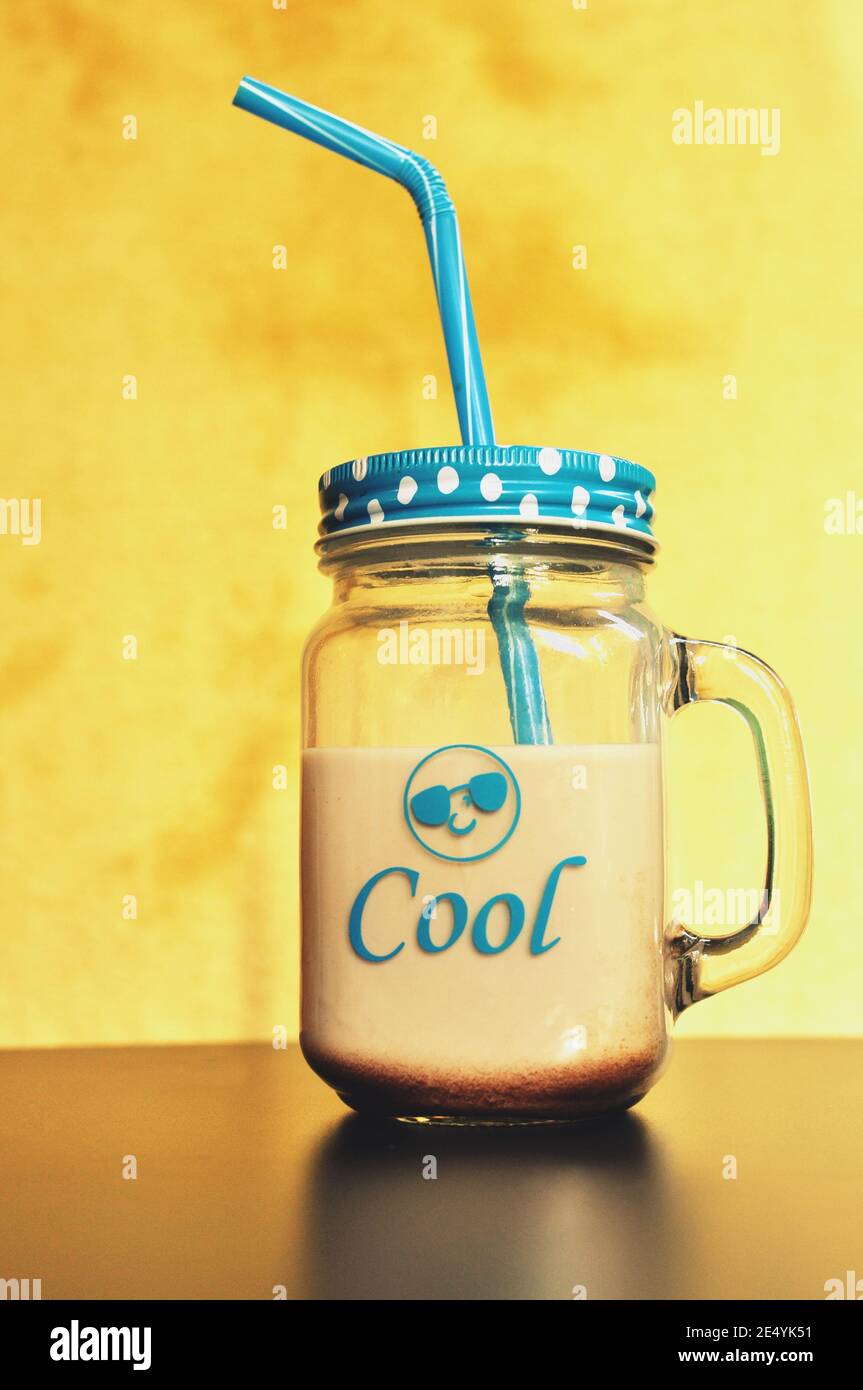 Kakao Milch trinken, in ein großes Glas mit Griff, Blau Deckel und auf gelben Hintergrund Stroh. Im vorderen ist cool und ein emoticon geschrieben. Ansicht von vorn Stockfoto