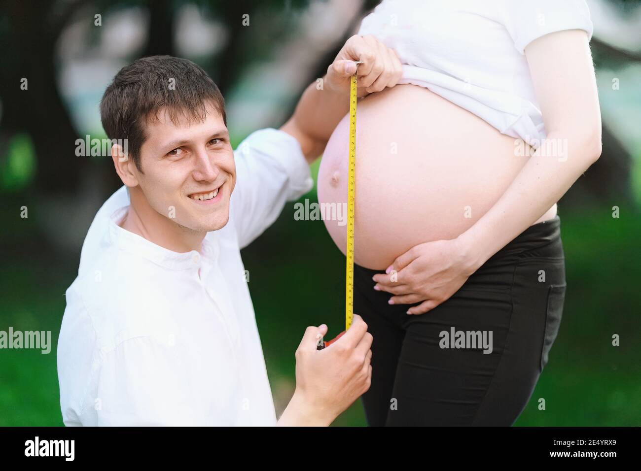 Ein junger Mann lächelt und misst den Magen seiner schwangeren Frau Mit einem Maßband in einem Sommerpark Stockfoto