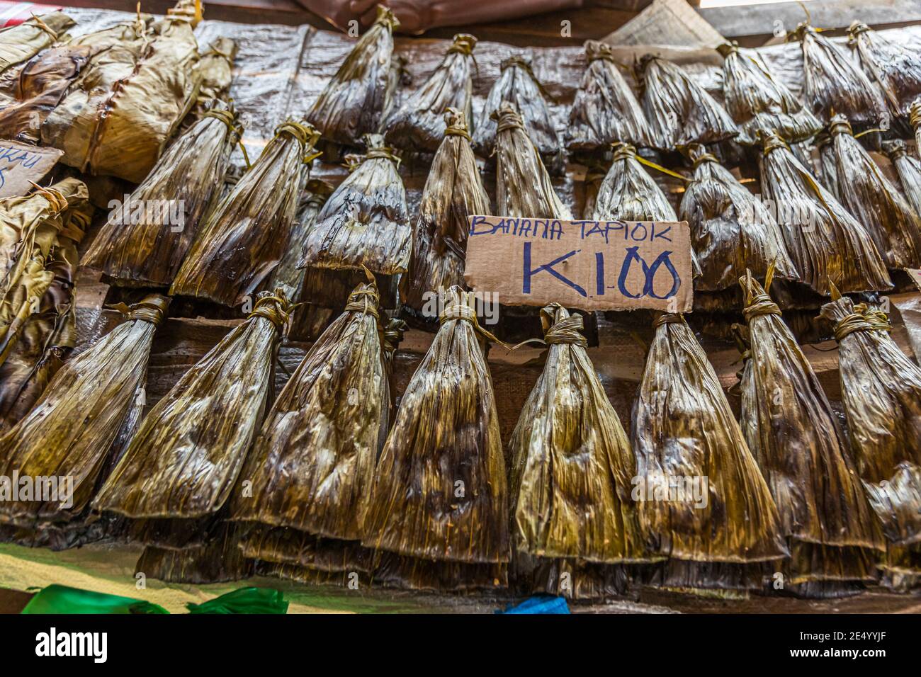 Getrocknete Bündel, in Blätter eingerollt, liegen in Reihen neben einem Schild mit der Aufschrift "Banana Tapiok K1.00" auf dem Markt in der Stadt Buka, Bougainville, Papua-Neuguinea. Stockfoto