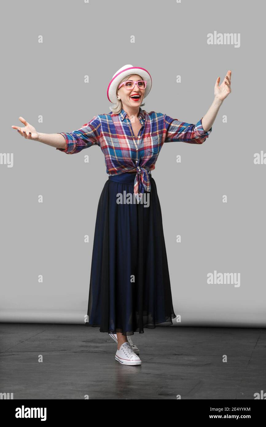 Volle Länge Portrait von glücklich moderne, elegante, reife Frau im lässigen Stil mit Hut und Brille stehend mit erhobenen Armen und überraschte Gesicht Stockfoto