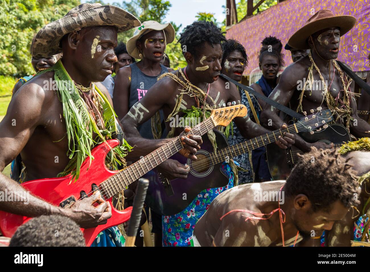 Musiker, die mit Palmenblätter geschmückt sind, spielen eine rote E-Gitarre und eine Akustikgitarre bei einem Sing-Sing-Dorf-Fest in Bougainville, Papua-Neuguinea, ein Mann mit einem Tarnhut steht neben ihnen. Stockfoto