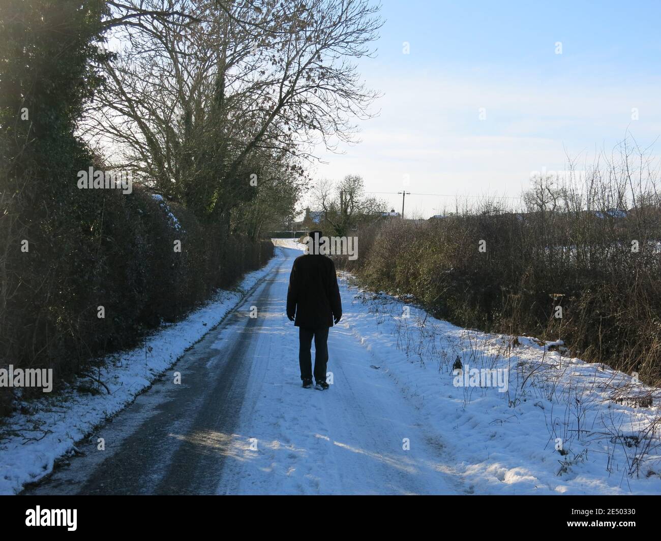 Rückansicht eines großen Mannes in dunkler Winterkleidung, der auf einer schneebedeckten englischen Landstraße unterwegs ist. Stockfoto