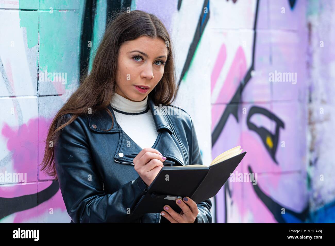 Eine junge Frau lehnt sich an eine mit Graffiti besprühte Wand Und macht Notizen in einem schwarzen Notizbuch Stockfoto