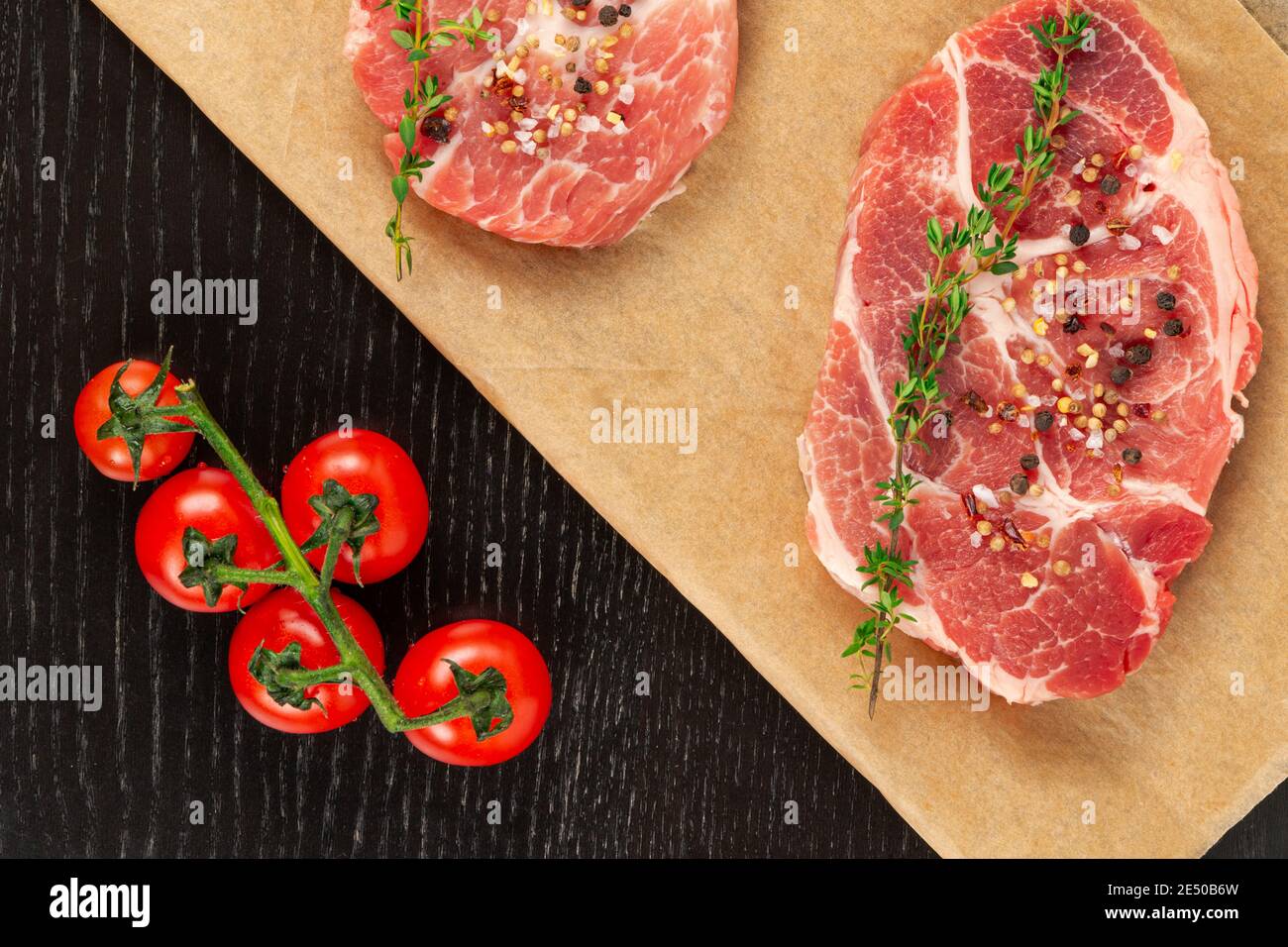 Frisch gehackte rohe Schweinesteaks mit Gewürzen und Thymian auf einem Blatt Pergament. Nahe reife rote Tomaten Stockfoto