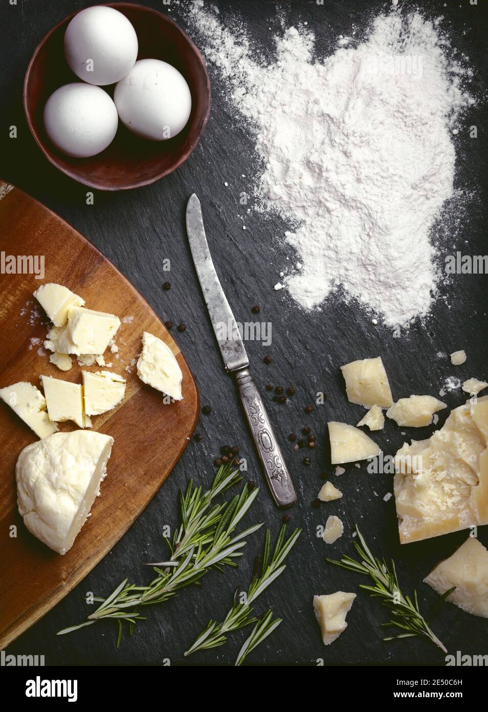 Backkäse Plätzchen Rezeptfoto leerer Essenshintergrund mit Parmesankäse, Butter, Eiern, Mehl, Rosmarin, Holzbrett und Vintage-Messer auf sla Stockfoto