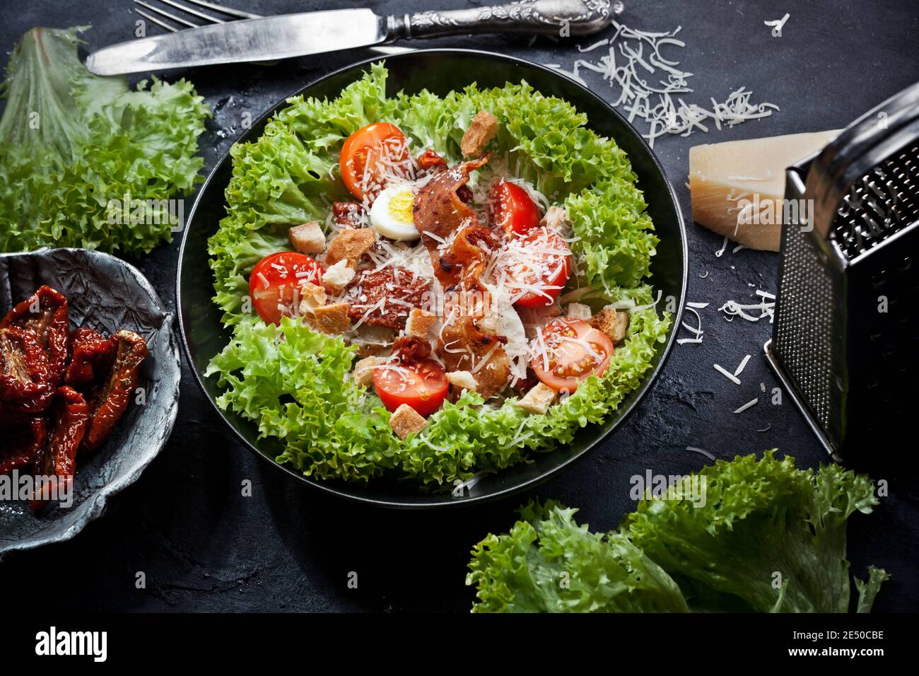 Flatlay Ansicht von frischem Caesar Salat mit Salatblättern, frischen und trockenen Kirschtomaten, geriebenem Parmesan Käse, Eier, Fleisch und Croutons, in einem schwarzen pla Stockfoto