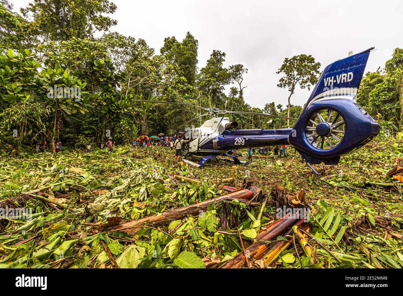 Der Heckrotor und der Ausleger des True-North-Hubschraubers, gekennzeichnet mit „VH-VRD“, heben sich im Vordergrund einer gerodeten Fläche im Regenwald von Bougainville, Papua-Neuguinea, ab, während sich Dorfbewohner mit Regenschirmen im Hintergrund um das Flugzeug versammelt haben. Stockfoto