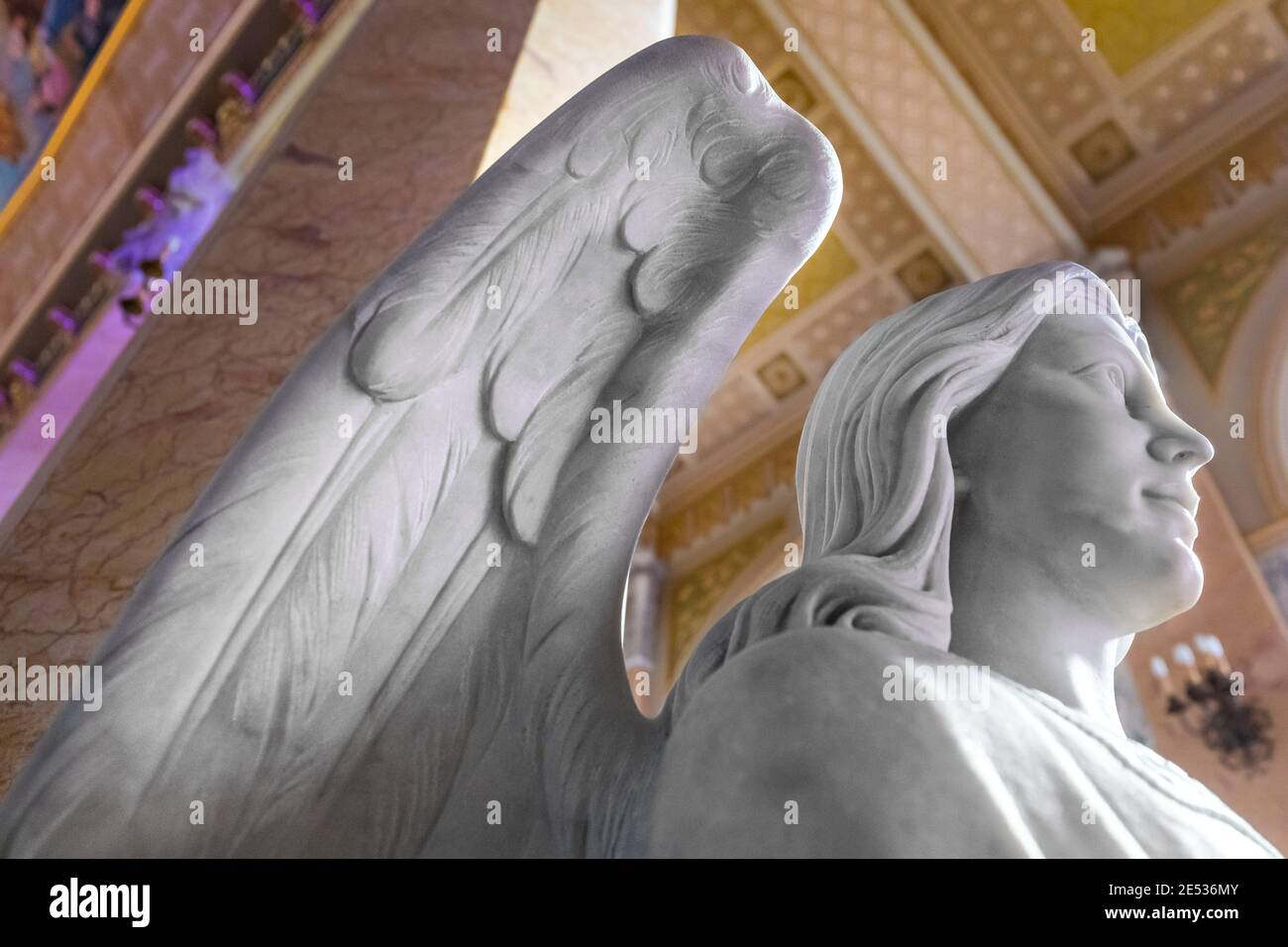 Nahaufnahme einer weißen Marmorstatue eines geflügelten Engel gegen das Kirchenschiff einer bunten Kirche Stockfoto