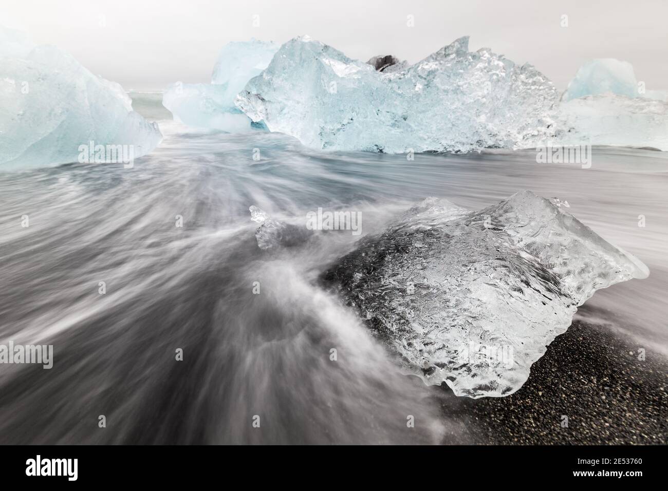 Nahaufnahme eines Eisblocks, der an einem schwarzen Ufer mit Wasser abschwellt und fließt, und hellblaue Eisberge im Hintergrund Stockfoto