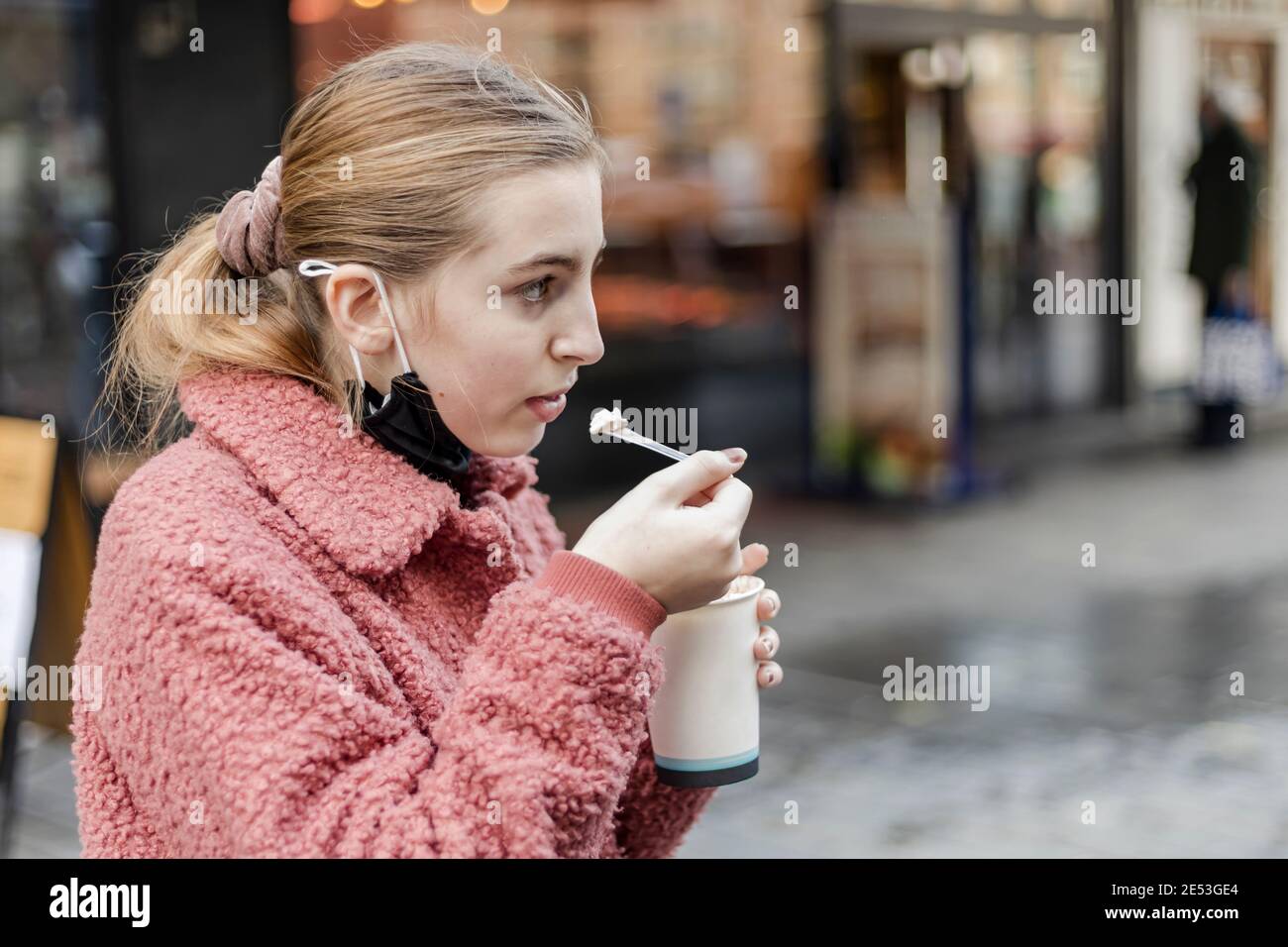 Blonde Teenager-Mädchen trägt eine Gesichtsmaske unter ihrem Kinn Essen Schlagsahne aus einer Papiertasse heiße Schokolade In einer städtischen Umgebung im Winter Stockfoto