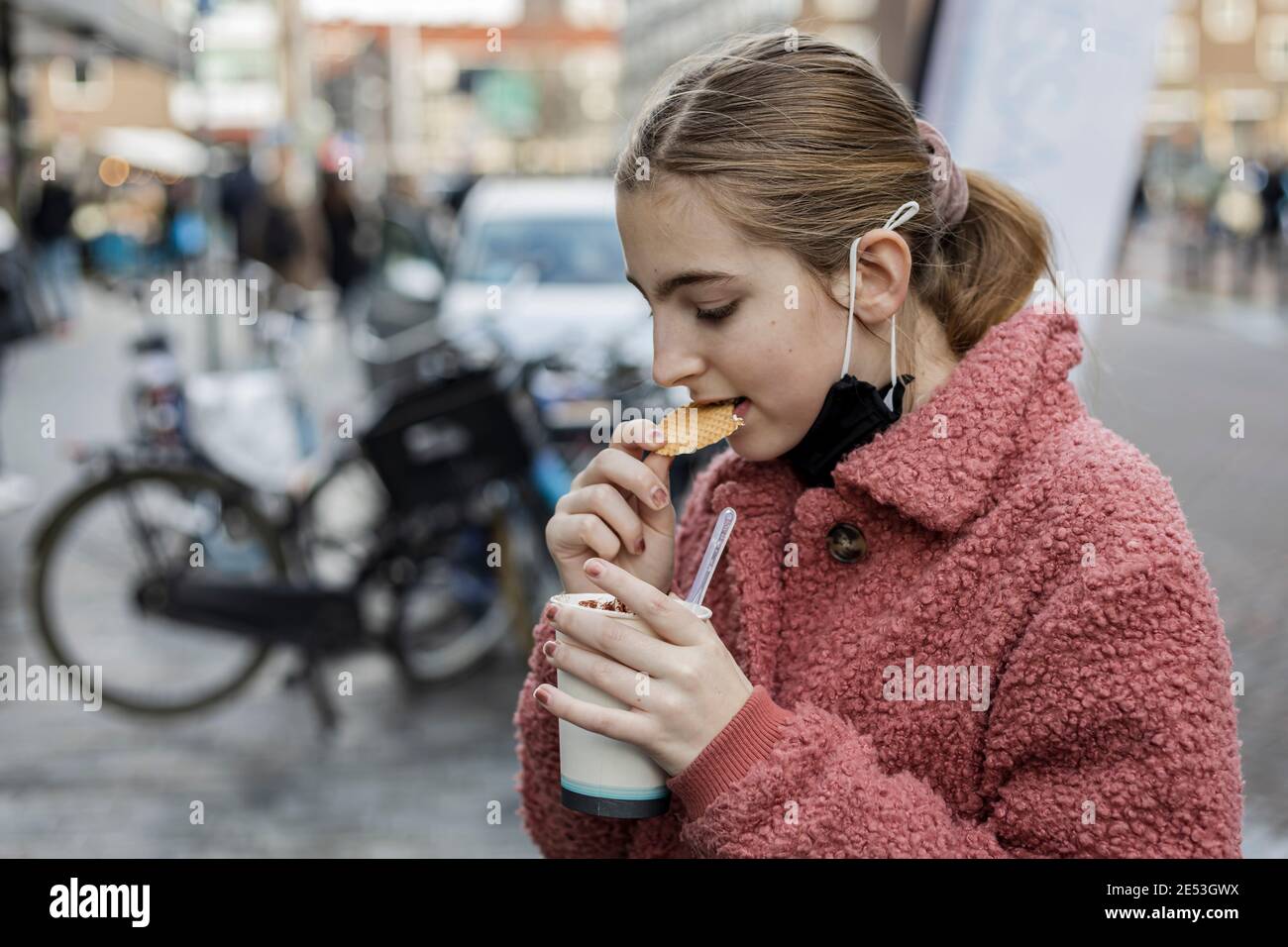 Junge blonde Mädchen trägt eine Gesichtsmaske unter ihrem Kinn, essen ein Cookie draußen in einer städtischen Umgebung Stockfoto