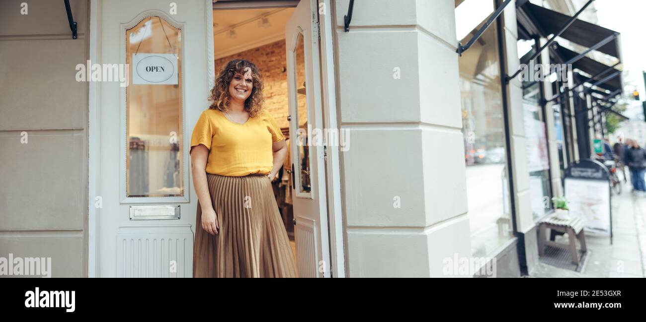 Portrait des Inhabers des Bekleidungsladens, der an der Tür steht und die Kamera anschaut. Schöne Frau, die einen Modehaus betreibt. Stockfoto