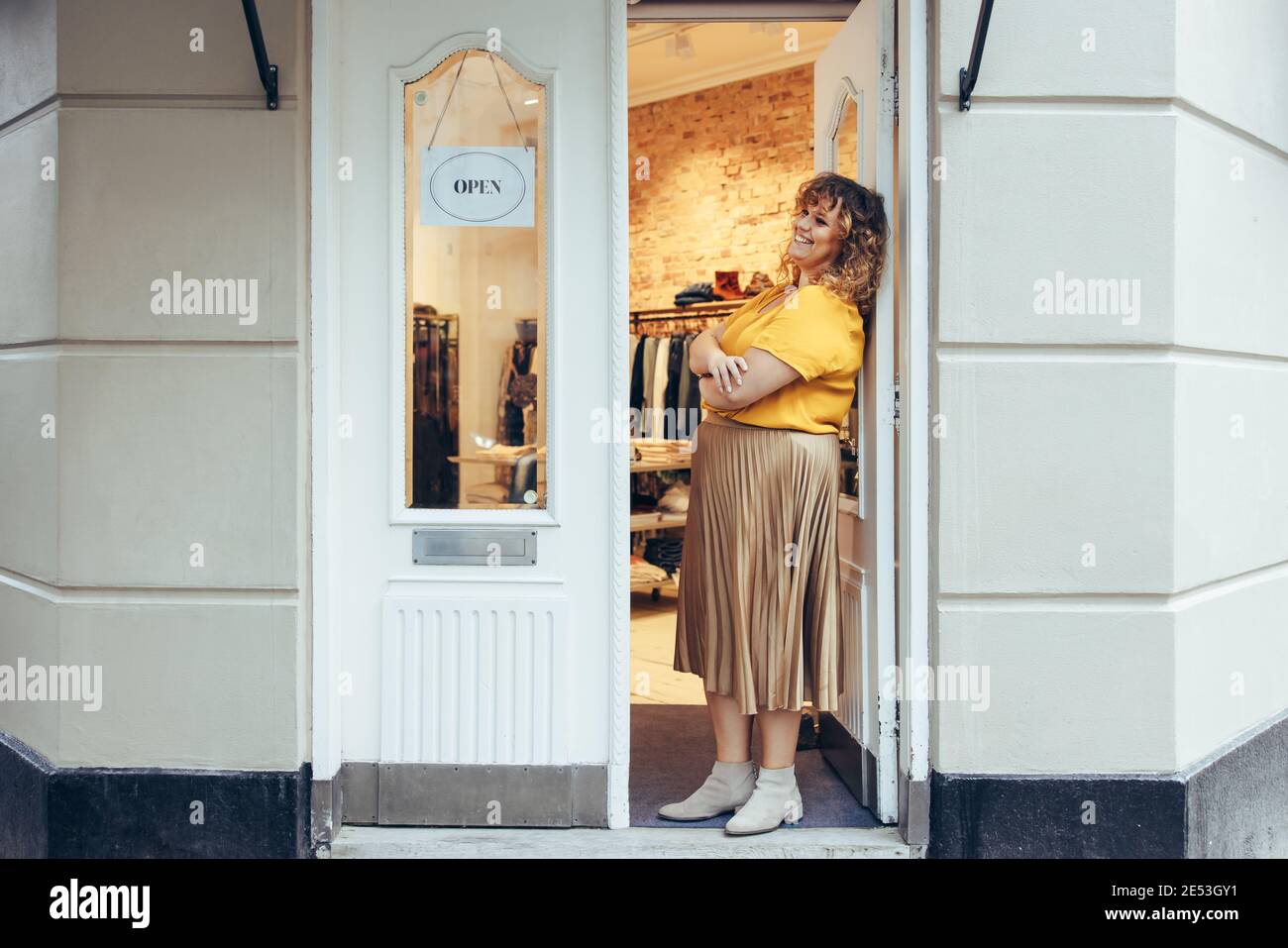 Besitzer eines Bekleidungsgeschäfts lehnt sich an die Eingangstür ihres Geschäfts. Geschäftsfrau steht vor der Tür einer Boutique. Stockfoto