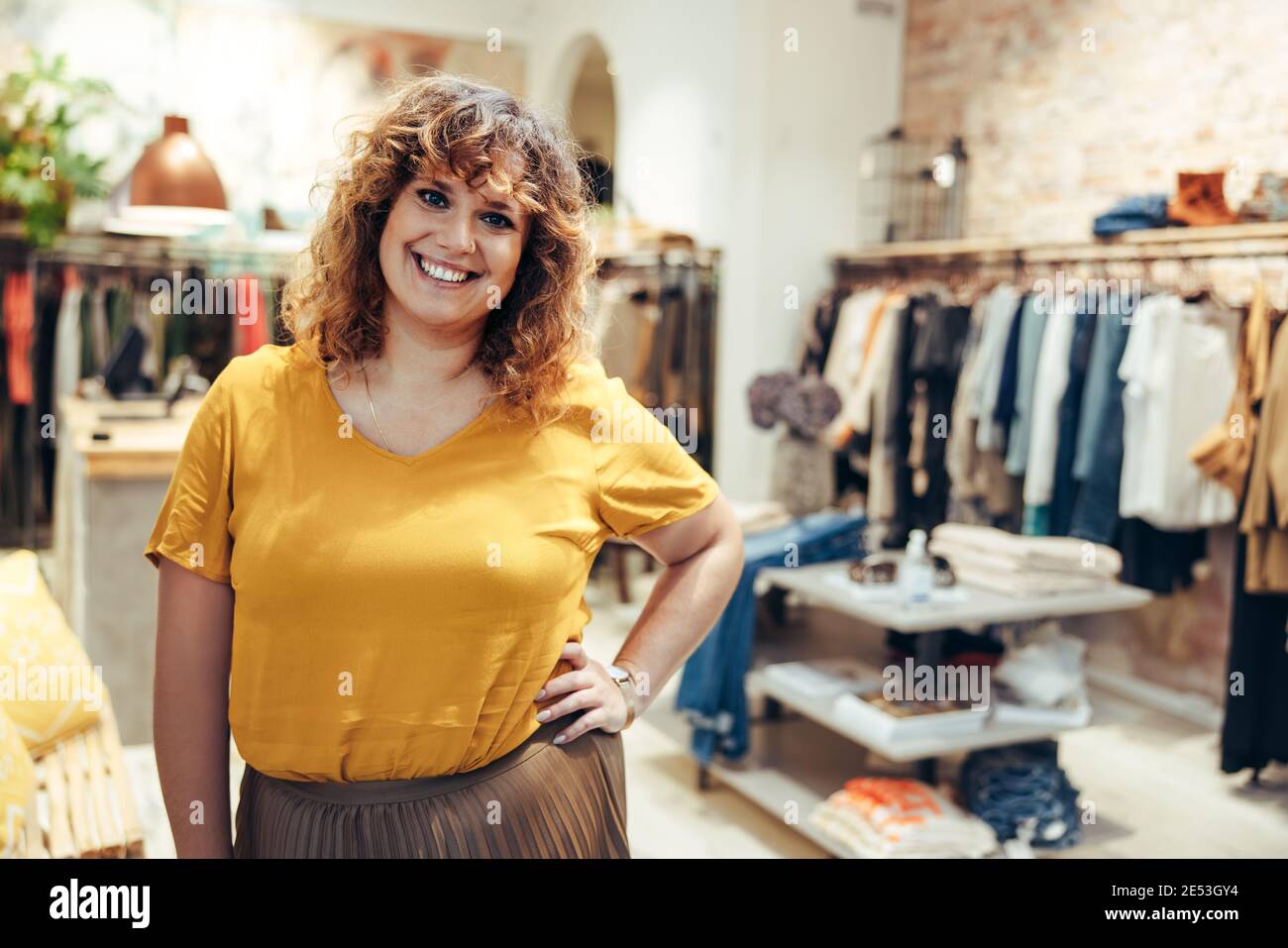 Portrait einer attraktiven Frau, die in einem Modehaus steht und mit der Hand auf der Hüfte auf die Kamera schaut. Glückliche reife Frau steht in einer Modeboutique. Stockfoto