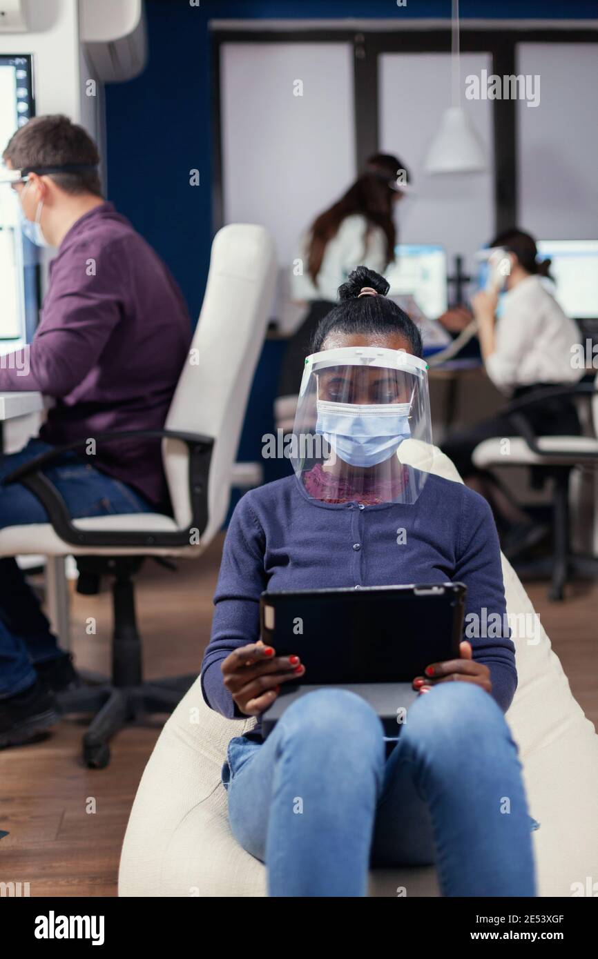 Afrikanischer Manager Notizen auf Tablet-pc trägt Gesichtsmaske gegen covid19 am Arbeitsplatz. Multiethnisches Geschäftsteam, das während der globalen Pandemie mit Coronavirus soziale Distanz respektiert. Stockfoto