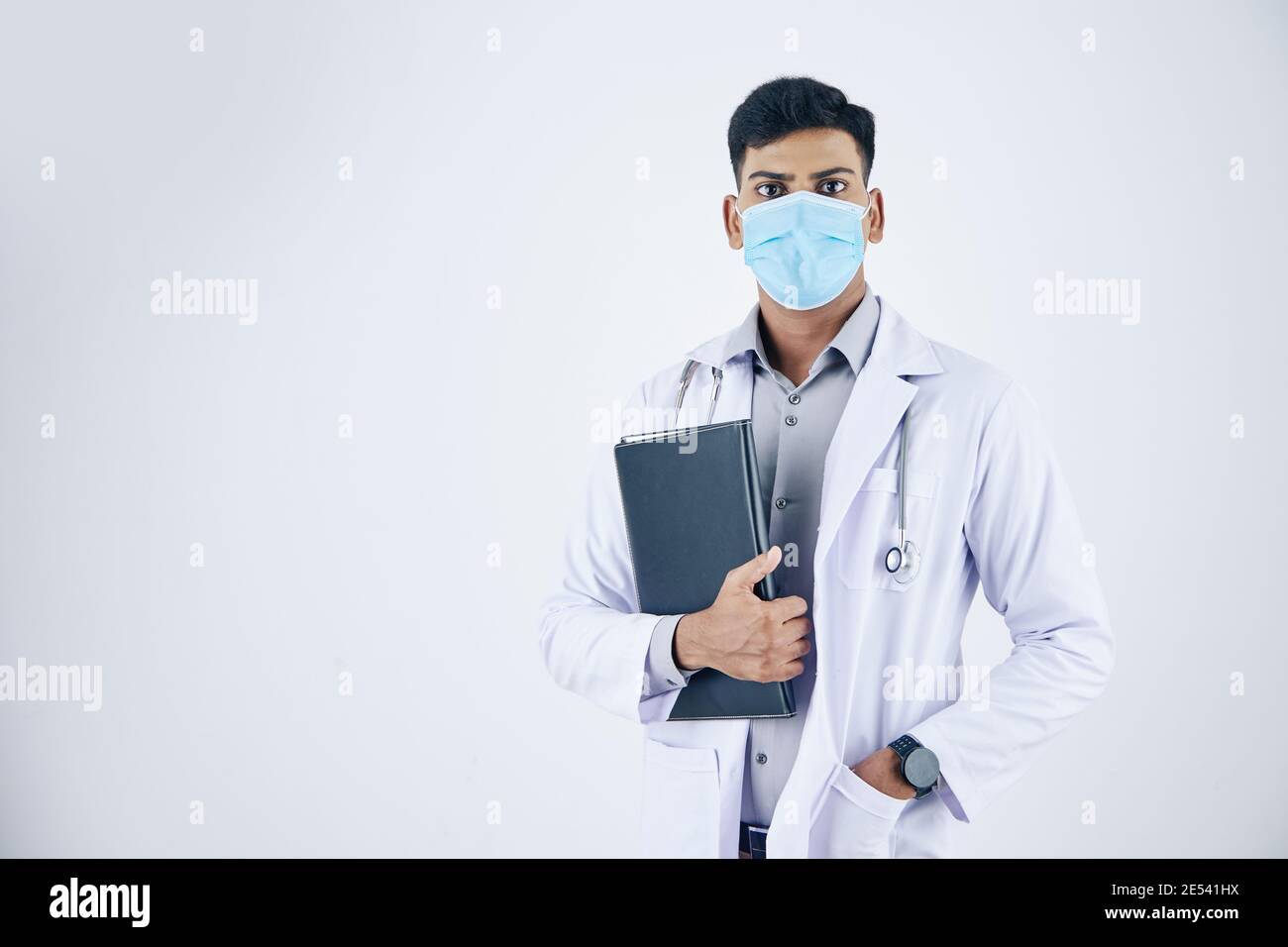 Porträt des jungen ernsten Arzt in medizinische Maske mit Leder Ordner mit Dokumenten und Blick auf die Kamera Stockfoto