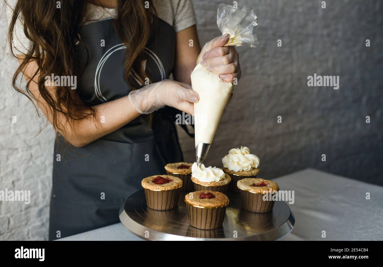 Das Mädchen kocht Cupcakes zu Hause. Hände eines Küchenchefs mit einer Tüte, die Sahne auf die Cupcakes quetschen Stockfoto